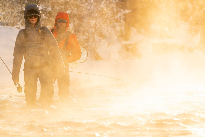 Two anglers wade through an icy river in moody late-day light
