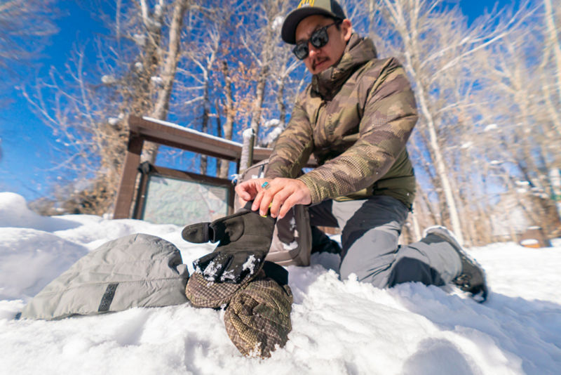 An angler kneeling in the snow puts on a pair of gloves that matches the camo of their hoodie