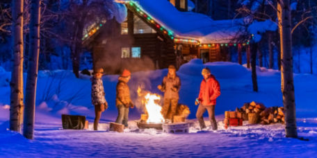 A group of people warming themselves by a campfire on a snowy night in front of a log cabin decorated with Christmas lights.
