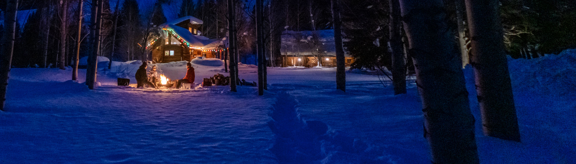 A lodge at night covered in a blanket of snow with holiday lights as trim in back of a group of people by a bonfire.