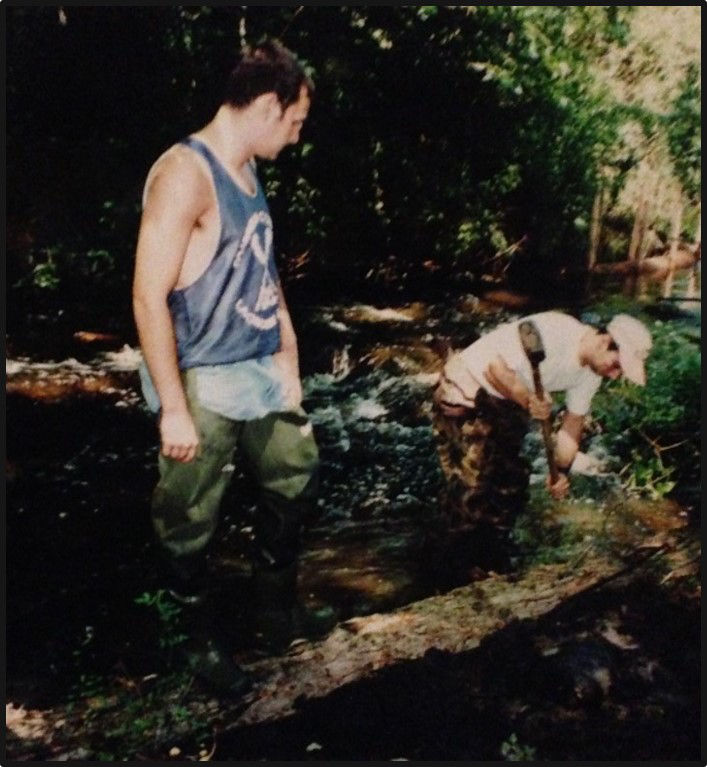 A man digs in the stream while another man watches.