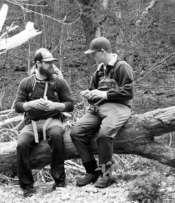 Black and white image of Jess and a friend in the woods wearing waders