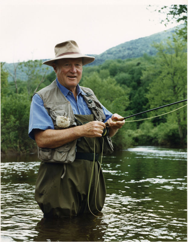 Leigh H Perkins, wearing fishing gear, stands hip-deep in water holding a fly rod.