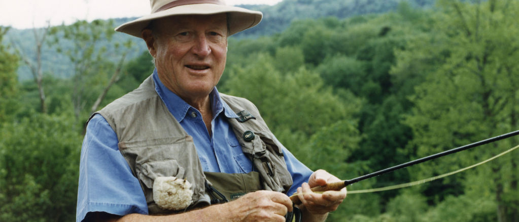 Leigh H Perkins, wearing fishing gear, stands hip-deep in water holding a fly rod.