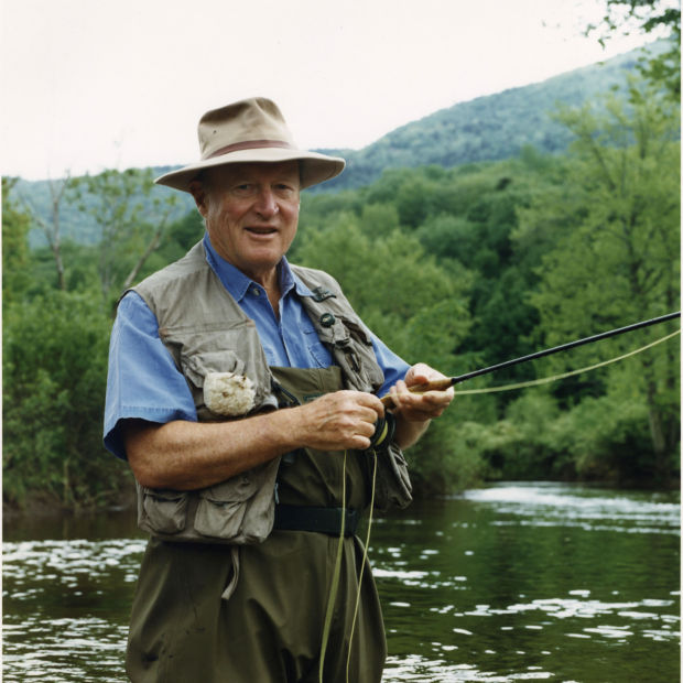 Leigh H Perkins, wearing fishing gear, stands hip-deep in water holding a fly rod.