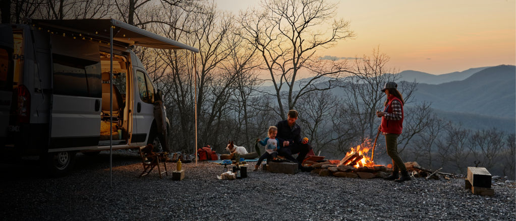 A family surrounding a campfire during sunset outside of their camper
