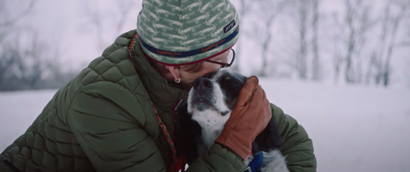 A woman in a green jacket and winter hat crouches down to hug a black and white dog in a snowy field surrounded by trees.