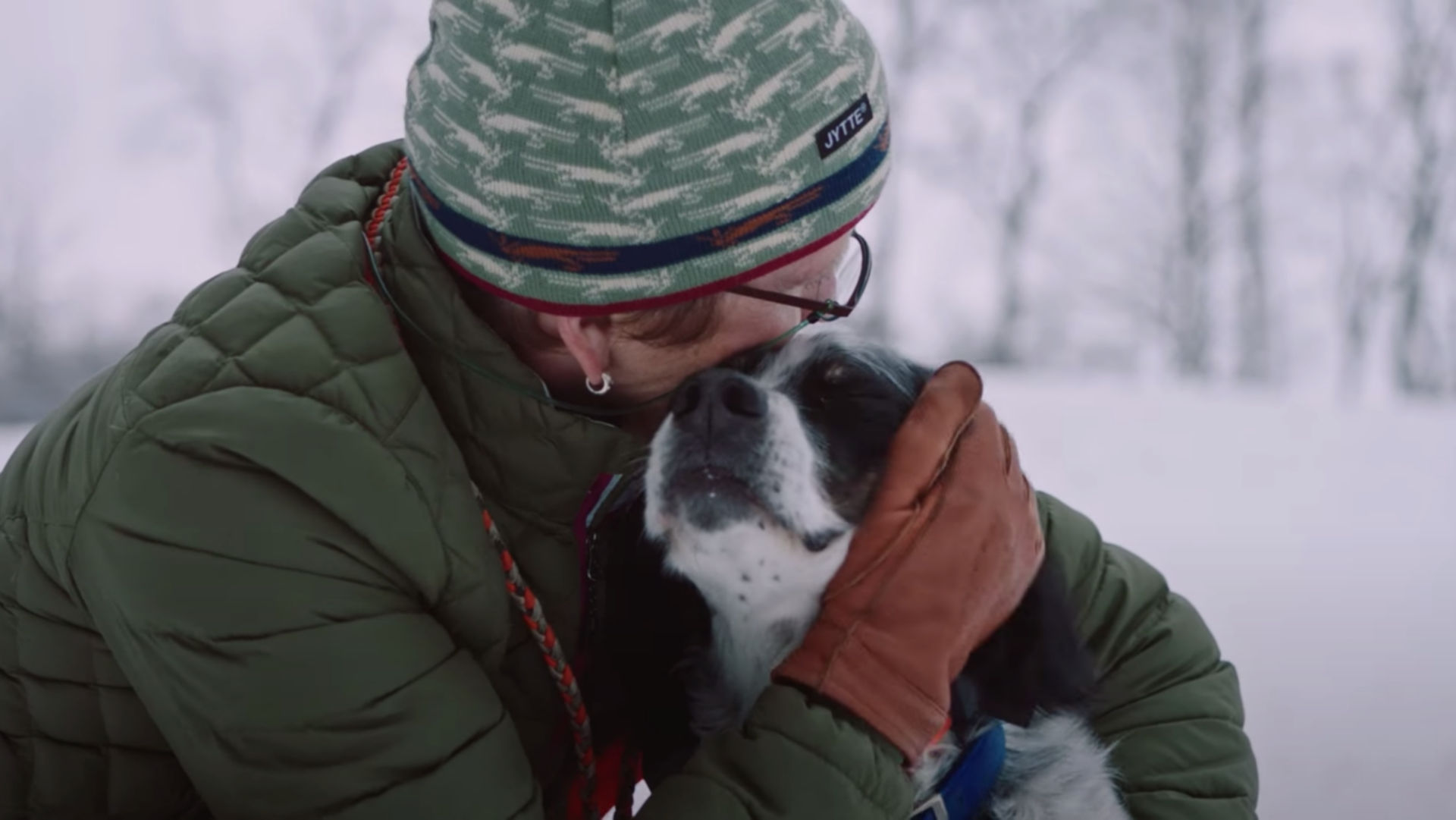 A woman in a green jacket and winter hat crouches down to hug a black and white dog in a snowy field surrounded by trees.
