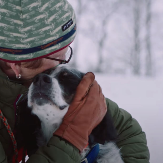 A woman in a green jacket and winter hat crouches down to hug a black and white dog in a snowy field surrounded by trees.