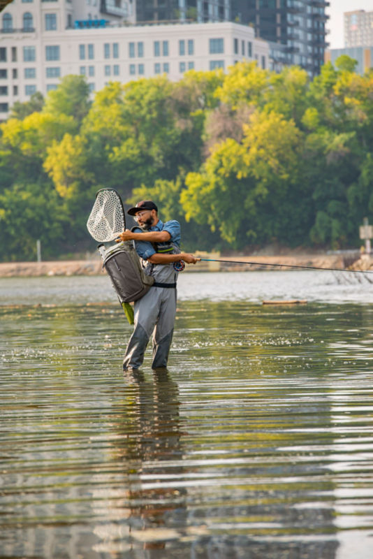 An angler reaches over his shoulder for his net from calf-deep in the water.