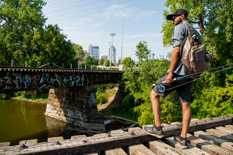 Man surveys urban river, rod in hand, from attop a bridge of railroad tracks.