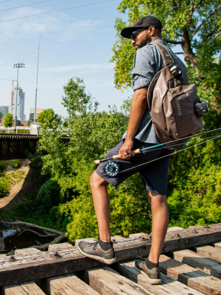 Man surveys urban river, rod in hand, from attop a bridge of railroad tracks.