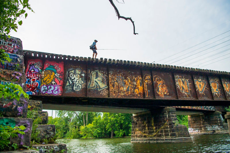 A view from below of an angler walking across a railroad bridge that is covered in colorful graffiti