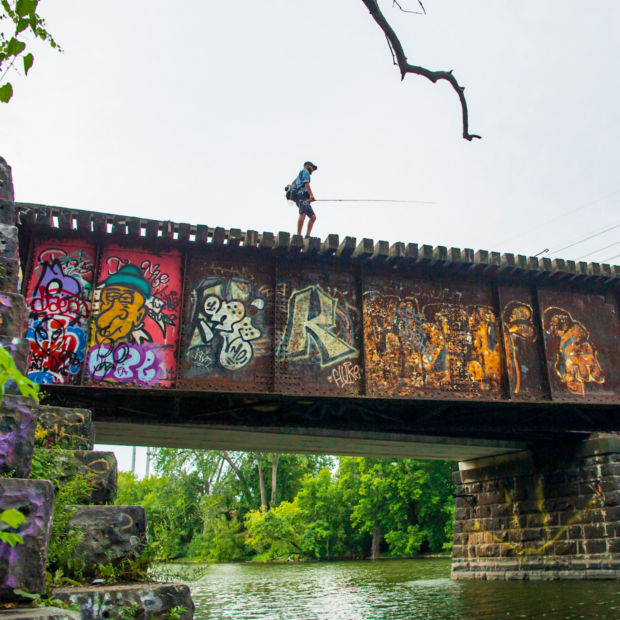 A view from below of an angler walking across a railroad bridge that is covered in colorful graffiti.