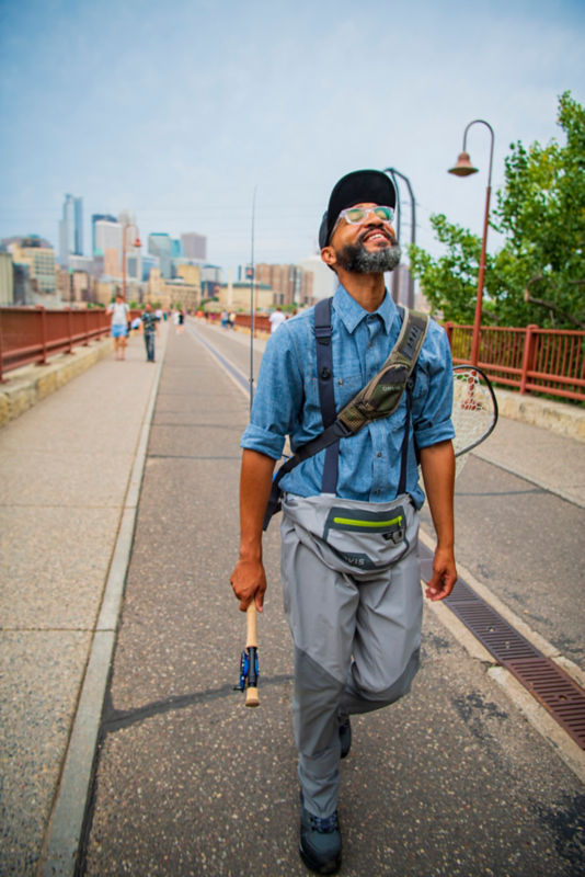 Ashley White smiling up at the sky as he walks a city bridge wearing full fly-fishing gear
