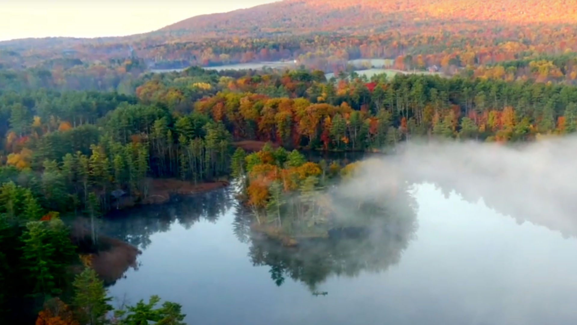 An aerial view of fog lifting off an autumn lake with hills in background