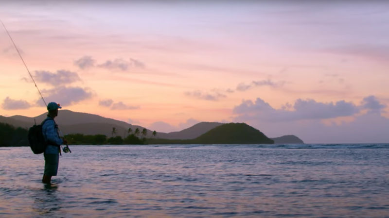 A sunset shot of a man standing in the water with mountains in the background