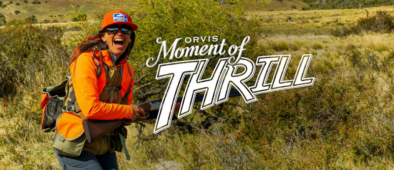 An excited woman wearing upland hunting gear in the field with the Moment of Thrill Logo