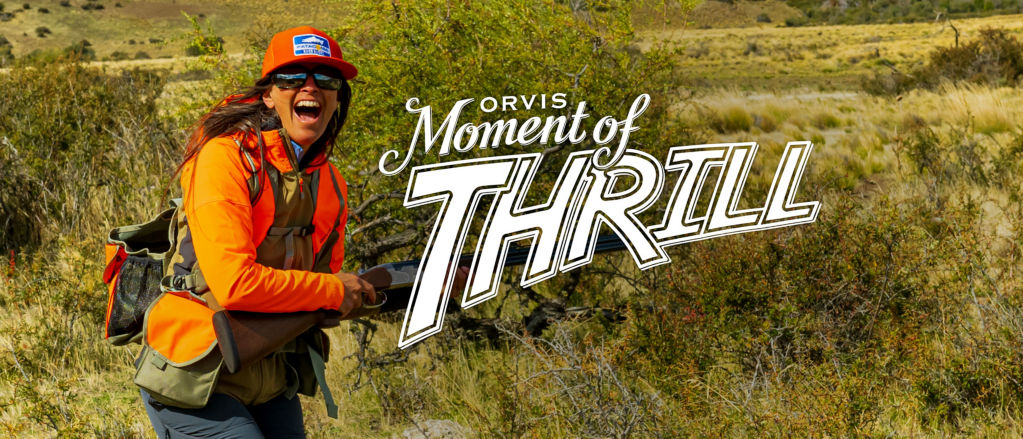 An excited woman wearing upland hunting gear in the field with the Moment of Thrill Logo