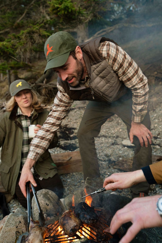 Charley Perkins in a green baseball cap and plaid shirt under a brown quilted vest cooks over a fire outside.