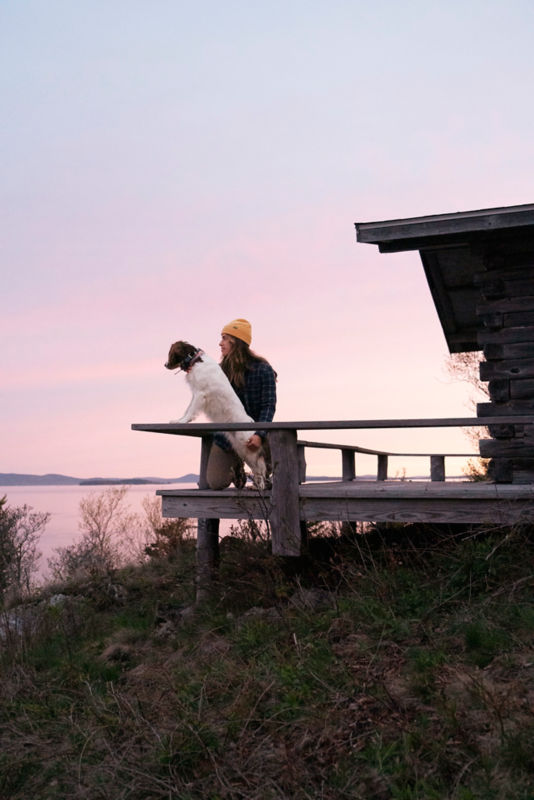 A woman sits with her dog on a wooden deck overlooking a calm lake at sunrise.