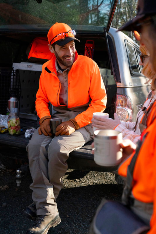 A hunter sitting on the tailgate of a truck wearing a blaze orange hat and jacket.