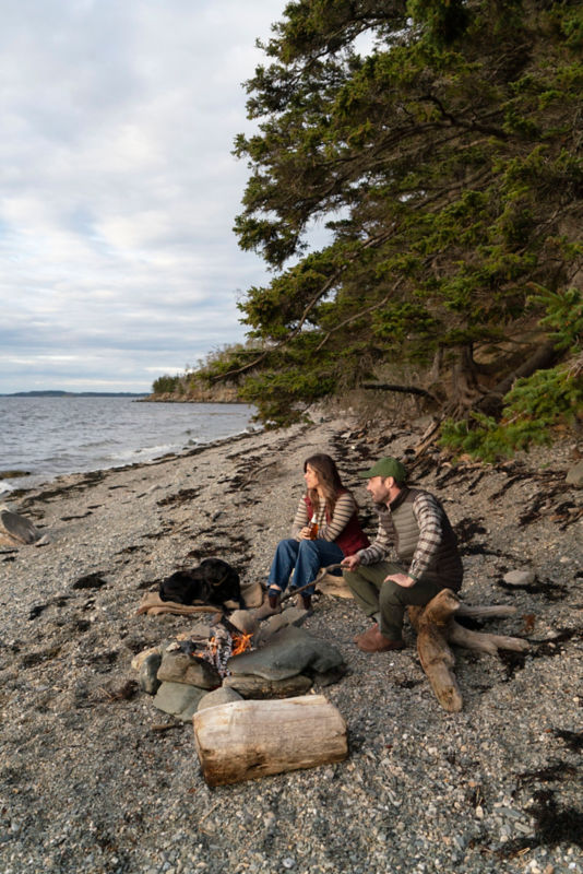 Two people dressed in casual clothing sit beside a small campfire on a rocky shoreline, facing the water.