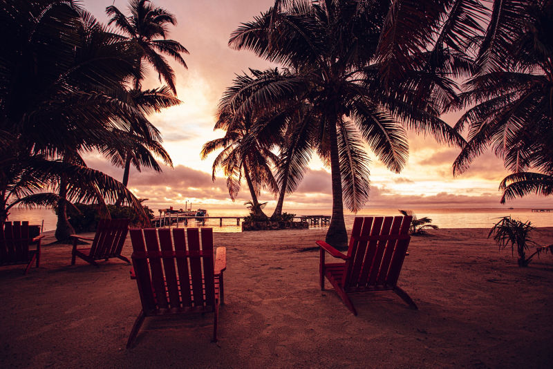 Several wooden chairs on a sandy beach looking out over a sunrise.