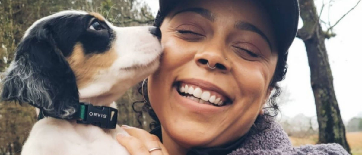 Melinda, wearing a hat and purple sweatshirt, smiles while her small white and black dog noses her cheek.