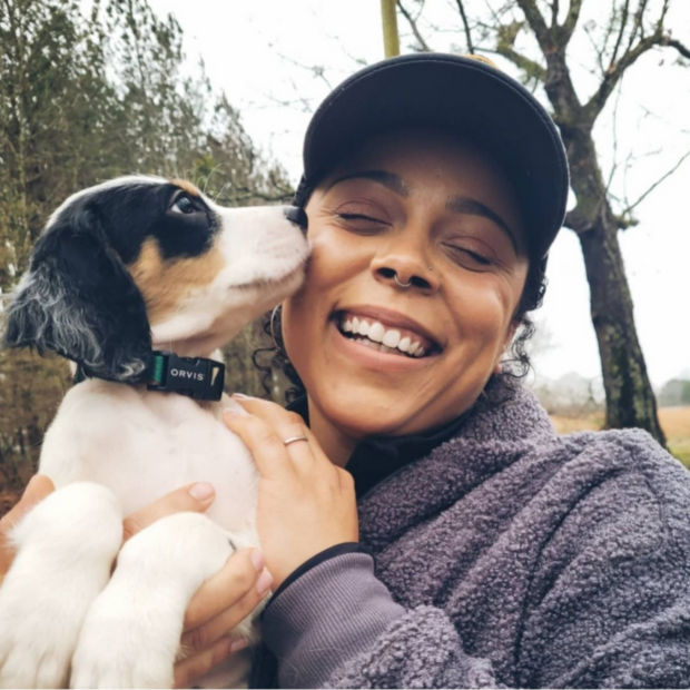 Melinda, wearing a hat and purple sweatshirt, smiles while her small white and black dog noses her cheek.