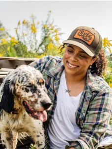 A smiling woman wearing a hat and flannel near her black and white dog