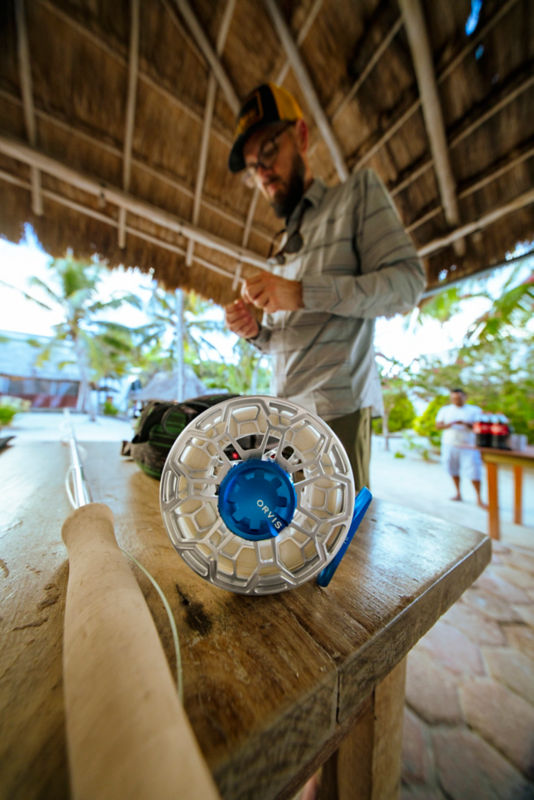 A silver Ratio fly reel with blue highlights sits cock-eyed on a wooden table next to a fly rod while an angler attaches a fly to a line.
