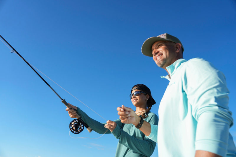 Benny guides his daughter in a cast with a fly rod under a bright blue sky.