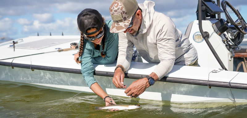 An angler and his daughter lean over the side of his boat, reaching into the water for a fish.