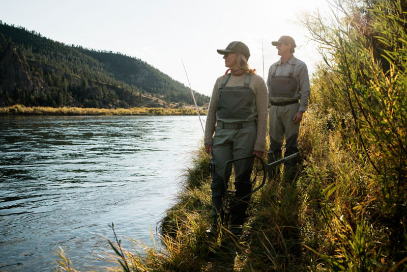 Two anglers walk an overgrown river bank to find the right spot for fishing.