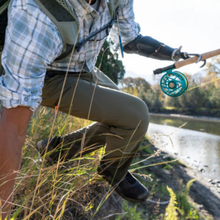 A man in a short-sleeve, blue plaid, button-down hops a riverbank to fly fish.