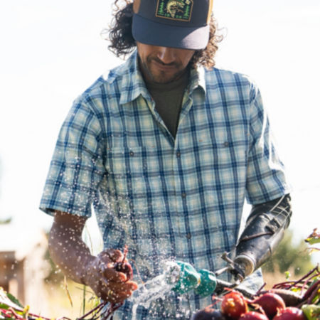 A man in a short-sleeve, blue, plaid, button-down washes a harvest of beets.