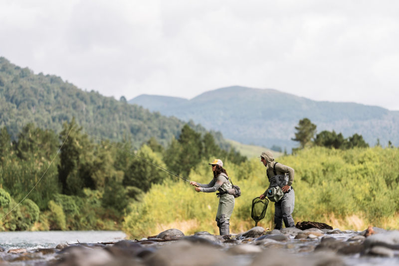 An angler reels in her fish while another angler prepares a net behind her.