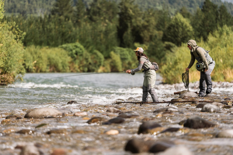 A guide stands behind an angler on a rocky spit.