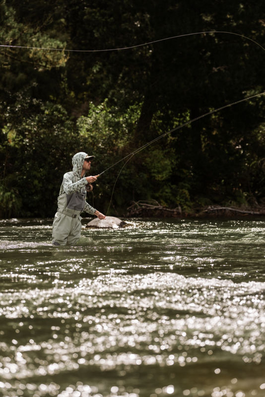An angler in waders and a light green camo sun protective hoodie.