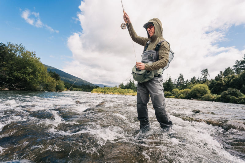 An angler in waders and an olive sun protective hoodie.