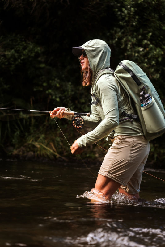 An angler leans into her fly rod while mending line from knee-deep in a river.