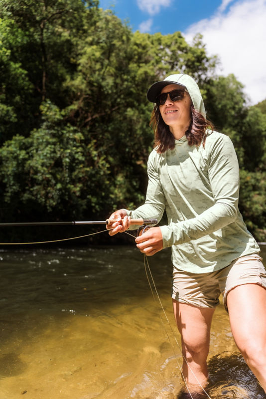 An angler wearing a light green sun protective hoodie while fishing in tea brown water.