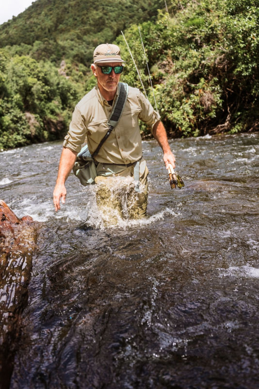 An angler wades thigh deep through a river wearing a tan Open Air Caster shirt.