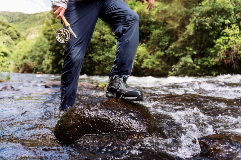 A waist-down view of a wet angler stepping up onto a large rock in a stream wearing navy Jackson Quick Dry Pants.