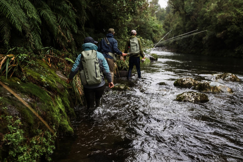 Three anglers loaded with packs and gear pick their way down the shallow part of a river.