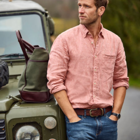 A man wearing jeans and a light red linen shirt leans against a jeep.