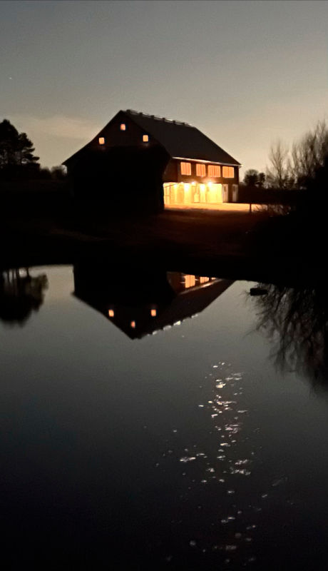 A view across a pond to a lit-up barn at night.