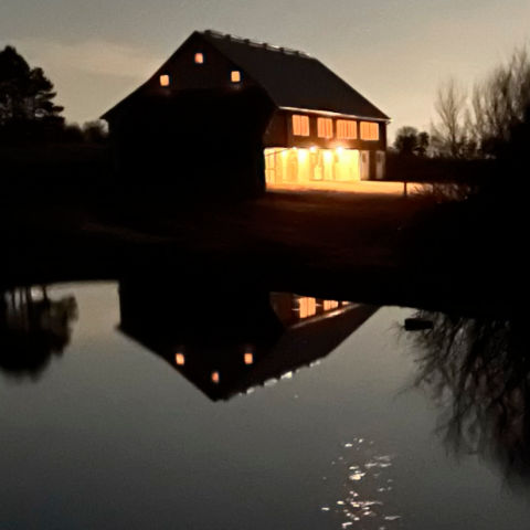 A view across a pond to a lit-up barn at night.
