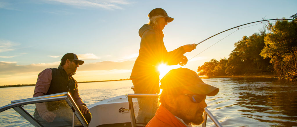 An angler casts her fly rod off a boat at sunrise.